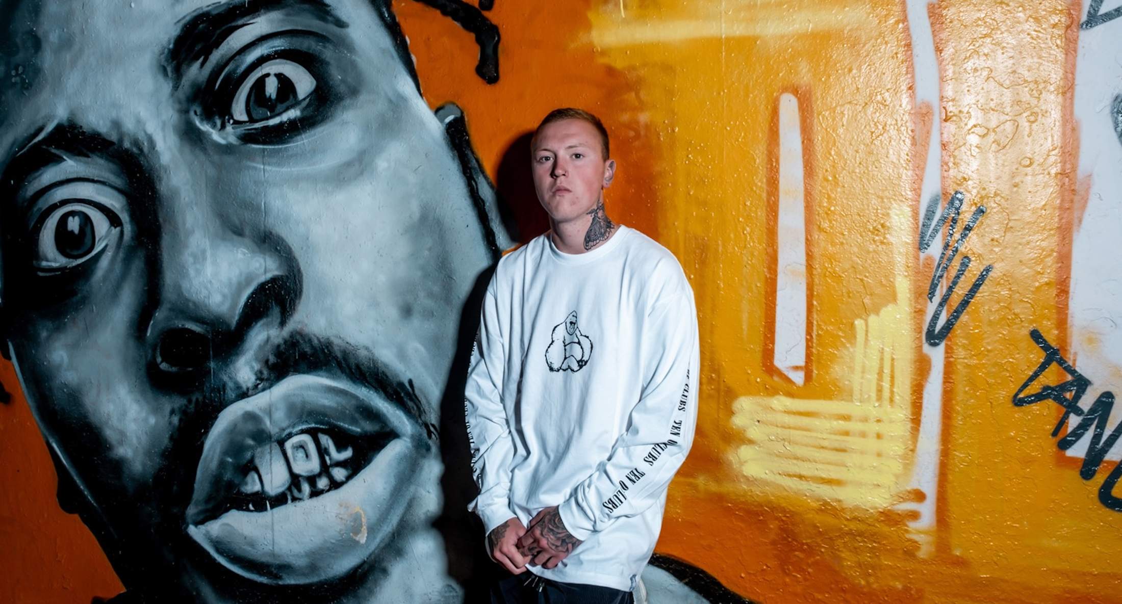 Man with tattoos in a TEN OF CLUBS Kong Long Sleeve Tee, stood in front of a wall of graffiti