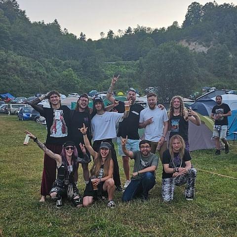 Group of people posing for a photo at an outdoor music festival, with trees and tents in the background.
