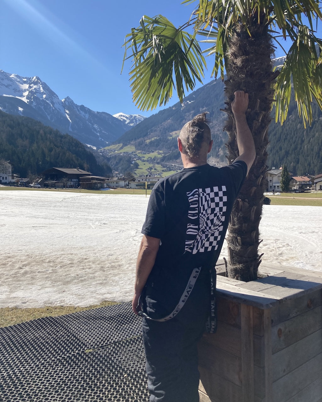 Man wearing Wave Tee by Ten of Clubs, standing by a palm tree with mountains and a lake in the background.