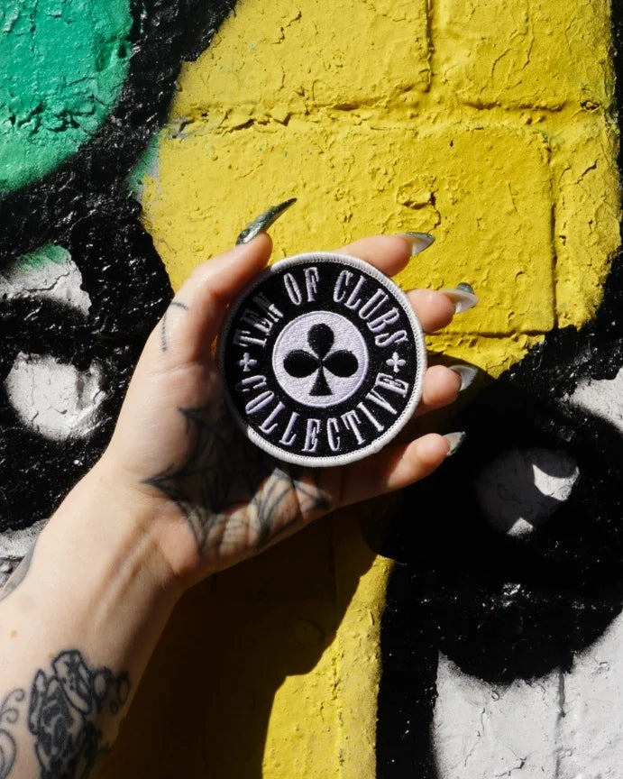 Black and white TEN OF CLUBS Collective embroidered patch held in a tattooed hand against a colourful graffiti wall.