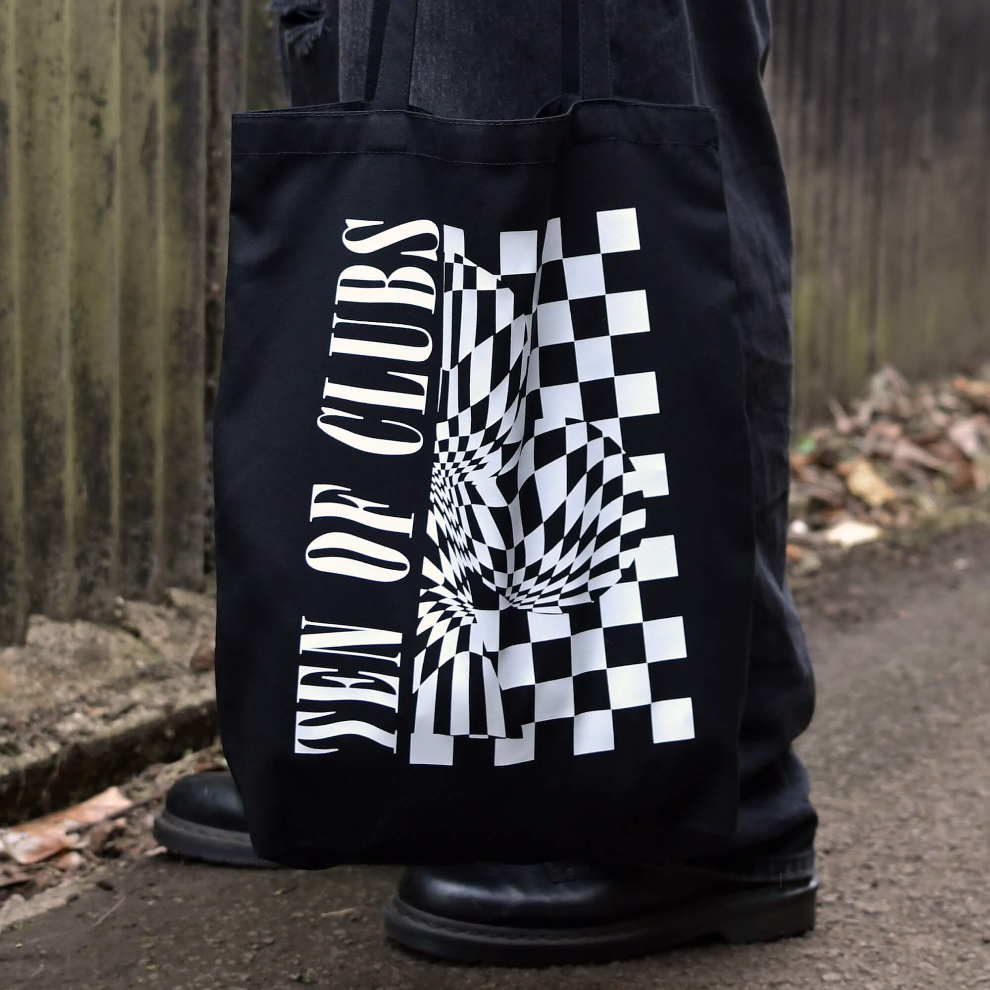 TEN OF CLUBS Wave Tote Bag in black with graphic artwork on the front, held by a person outdoors.