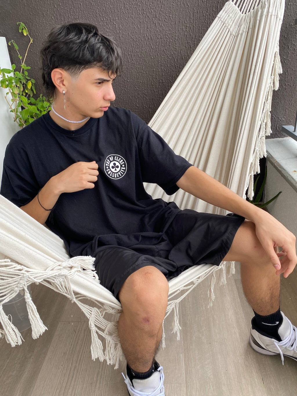 Young man wearing black Gunslinger Tee by Ten of Clubs, sitting on a white hammock indoors