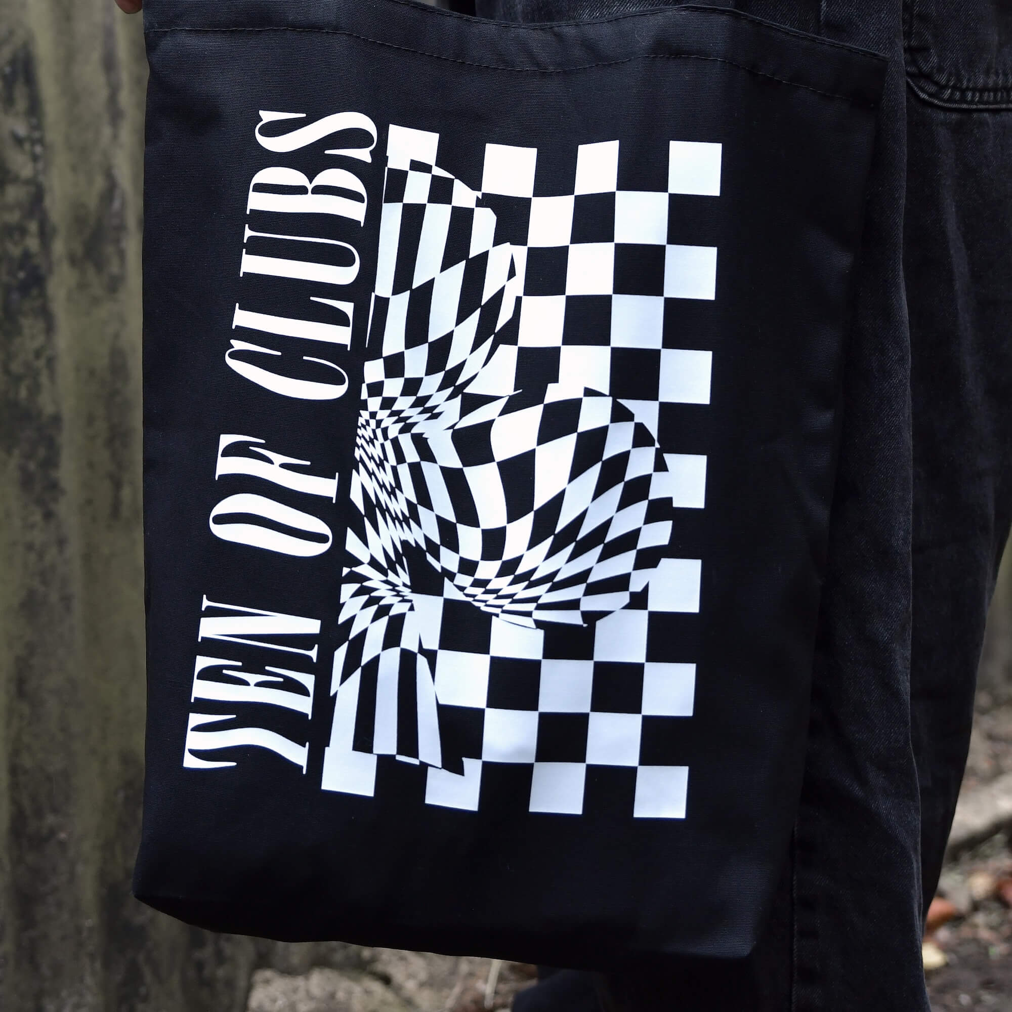 TEN OF CLUBS Wave Tote Bag in black with graphic artwork on the front, held by a person outdoors.