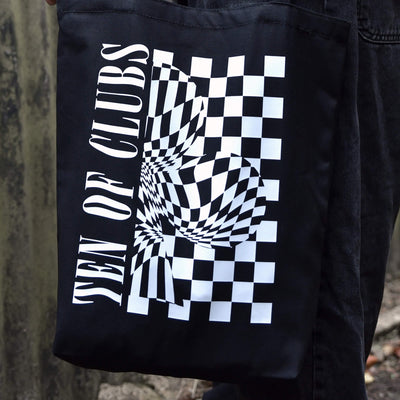 TEN OF CLUBS Wave Tote Bag in black with graphic artwork on the front, held by a person outdoors.